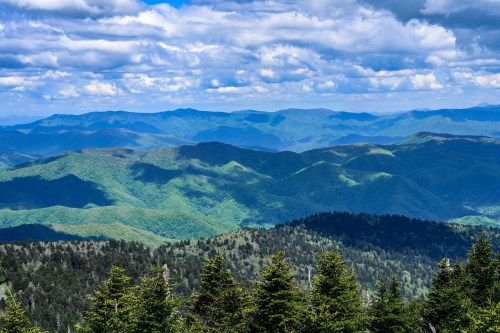 A panoramic view of lush green mountains under a partly cloudy sky, showcasing rolling hills and distant peaks.
