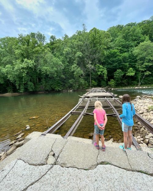 Two children stand on a rocky riverbank, looking at an old, partially collapsed wooden bridge surrounded by lush greenery.