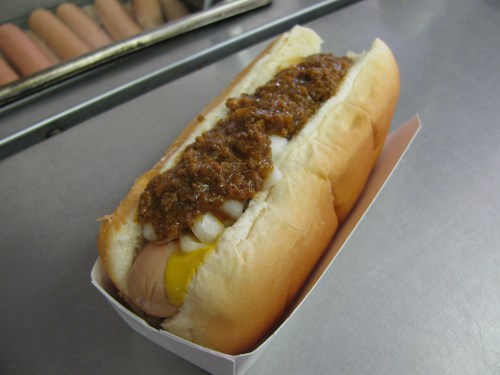 A hot dog in a bun topped with chili and diced onions, served in a paper tray.