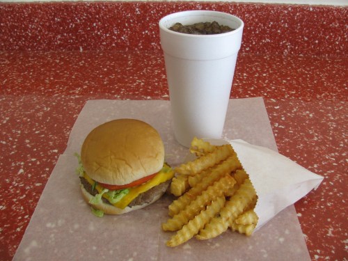 A cheeseburger with lettuce and tomato, crinkle-cut fries in a paper bag, and a large drink in a foam cup on a red countertop.