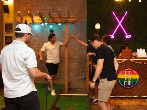 A group of friends plays mini-golf indoors, with colorful lights and a vibrant atmosphere in the background.