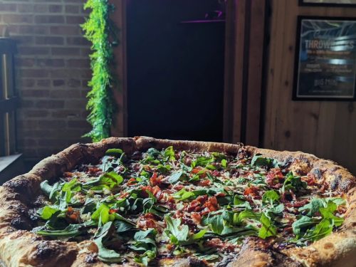 A close-up of a pizza topped with fresh greens and bacon, set against a rustic wooden background.
