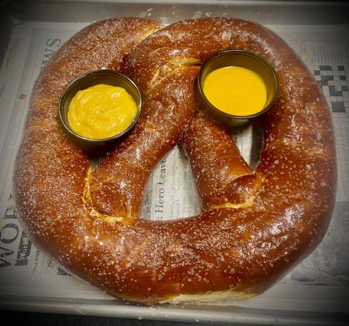 A large, soft pretzel sprinkled with salt, served with two bowls of mustard and cheese dip on a tray.