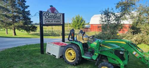 A green tractor parked by a sign for Barthel Fruit Farm, with a crate of apples nearby and orchards in the background.