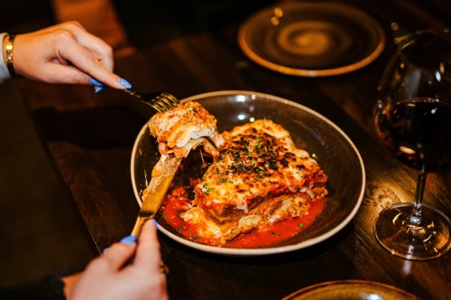 A person cuts into a serving of lasagna topped with herbs, served on a dark plate with a glass of red wine nearby.