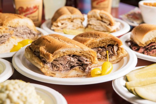 A close-up of a roast beef sandwich on a plate, surrounded by other sandwiches and sides, with pickles and peppers.