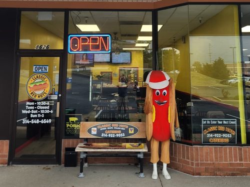 A hot dog mascot stands outside a restaurant with an "OPEN" sign, next to a bench and menu boards.