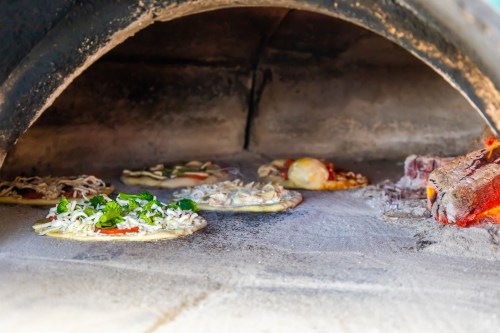 Pizzas baking in a wood-fired oven, with various toppings visible and glowing embers in the foreground.