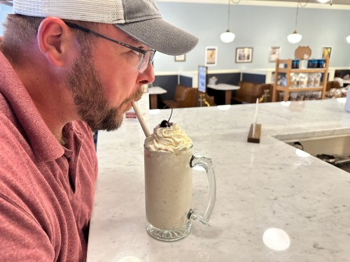 A man with a beard sips a milkshake topped with whipped cream and a cherry at a diner counter.