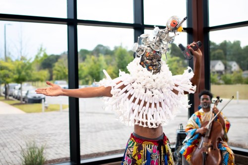 A performer in a creative costume with a unique headpiece gestures while another musician plays a cello in the background.