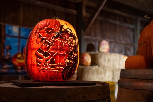 A carved pumpkin featuring a skeleton couple in a kiss, illuminated in a rustic barn setting.