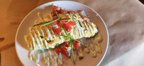 A plated omelette topped with diced tomatoes, drizzled with sauce, and garnished with chives on a white dish.
