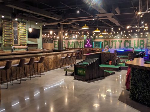 Interior of a mini putt club featuring a bar, putting greens, and vibrant decor with string lights.