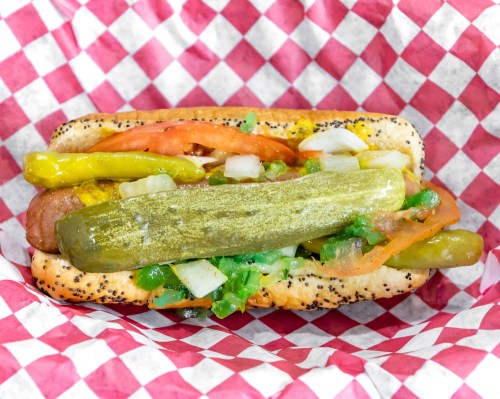 A hot dog topped with pickles, tomatoes, onions, and peppers, served in a checkered paper wrapper.