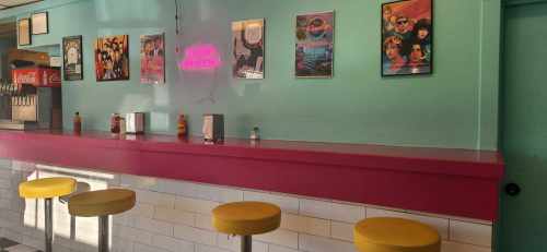 A colorful diner interior with a pink counter, yellow stools, and retro posters on the turquoise wall.