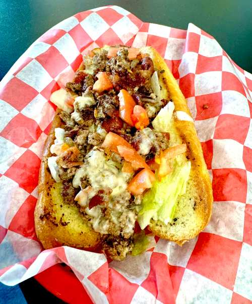 A loaded sandwich with ground meat, cheese, lettuce, and tomatoes on a toasted bun, served on a red and white checkered paper.