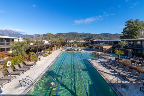A serene pool area with lanes for swimming, surrounded by lounge chairs and mountains in the background under a clear blue sky.