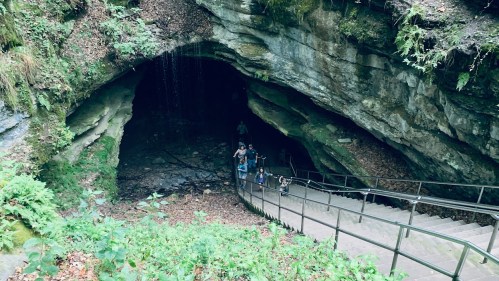 A group of people stands at the entrance of a cave, with a staircase leading down into the dark interior.