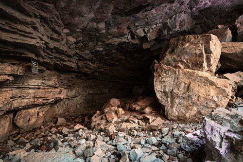 A rocky cave interior with large boulders and scattered stones on the ground, illuminated by soft light.