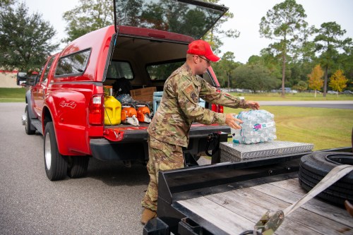 A soldier in camouflage unloads bottled water from a red pickup truck in a parking lot.