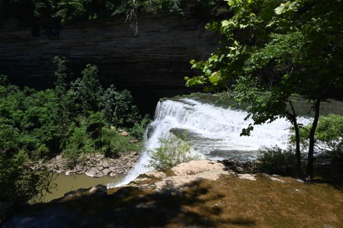 A scenic waterfall cascading over rocks, surrounded by lush green trees and a rocky landscape.