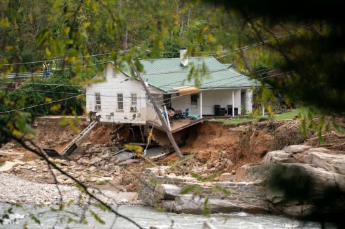 A house partially collapsed due to erosion, with exposed foundations and surrounding debris near a river.