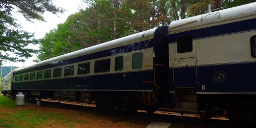 A blue and white train car parked among trees, with a green hillside in the background.