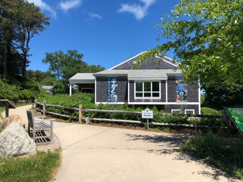A small aquarium building surrounded by greenery, with a clear blue sky above and a pathway leading to it.