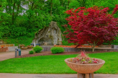 A serene garden with a stone grotto, vibrant red tree, and seating area surrounded by lush greenery.