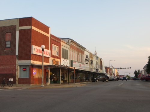 A quiet street scene featuring brick buildings, shops, and a setting sun in a small town.