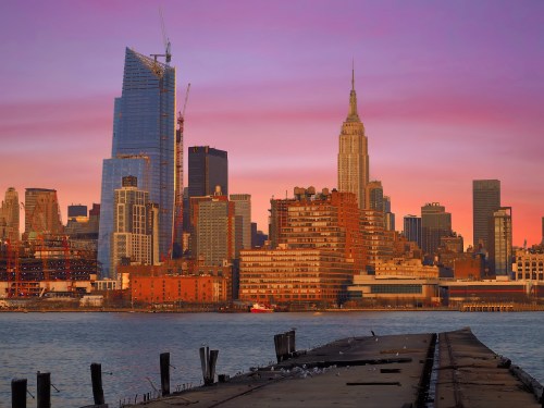 City skyline at sunset featuring the Empire State Building and modern skyscrapers, with a waterfront view.