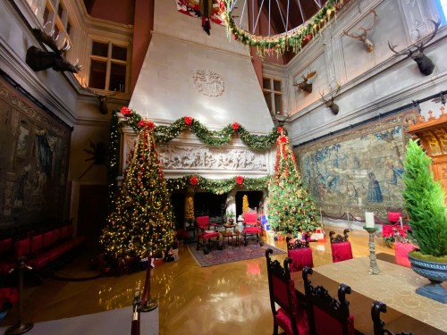 A festive room decorated for Christmas with two trees, garlands, and a grand fireplace, surrounded by elegant furnishings.