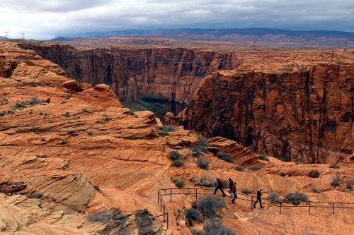 A scenic view of a canyon with steep red rock formations and a river below, featuring hikers on a winding path.