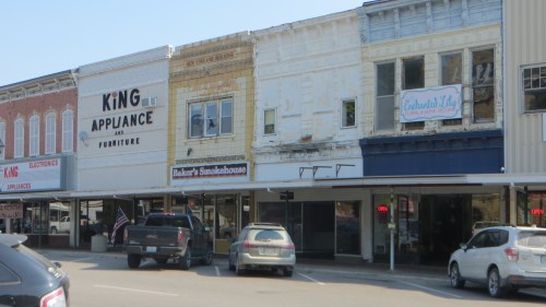 Historic storefronts line a street, featuring King Appliance, Baker's Smokehouse, and Celebrated Joy, with parked cars in view.