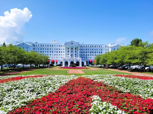 A grand white hotel building with a blue sky, surrounded by vibrant flower beds in red and white.