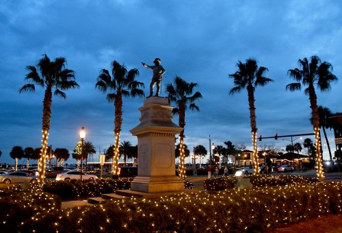 A statue stands among palm trees, adorned with lights, against a twilight sky with clouds.