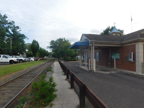 A train station with a brick building, a blue awning, and a nearby railroad track lined with trees and parked cars.