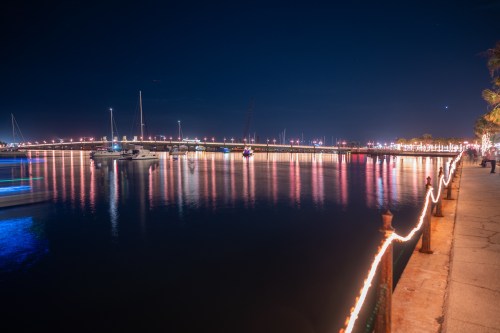A serene night scene of a waterfront with boats, lights reflecting on the water, and a bridge in the background.