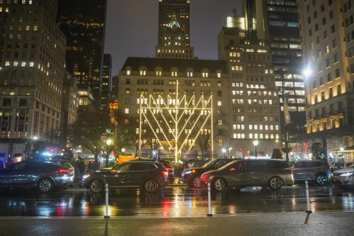 City street at night with illuminated building and traffic, reflecting lights on wet pavement.