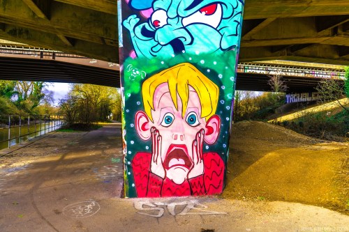 Colorful graffiti under a bridge features a shocked character with wide eyes and hands on their face, surrounded by vibrant designs.