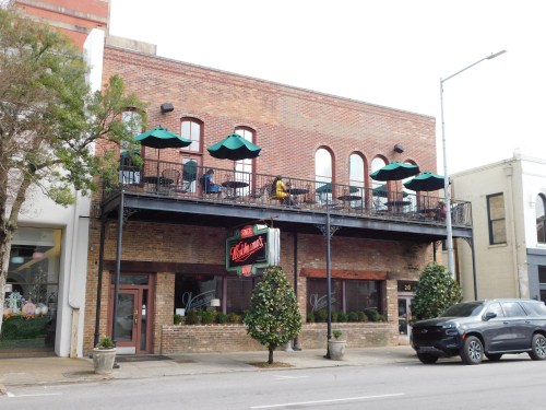 A brick building with a balcony and green umbrellas, featuring a restaurant sign and outdoor seating.