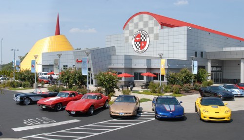 A lineup of classic and modern Corvettes parked in front of a sleek, modern building with a yellow cone-shaped roof.