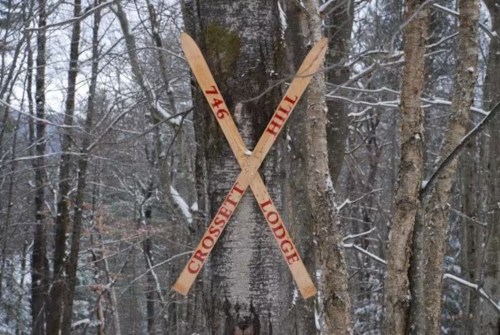 Two crossed wooden skis on a tree, marked with "746" and "Crossfit Hill Lodge," surrounded by snow-covered trees.