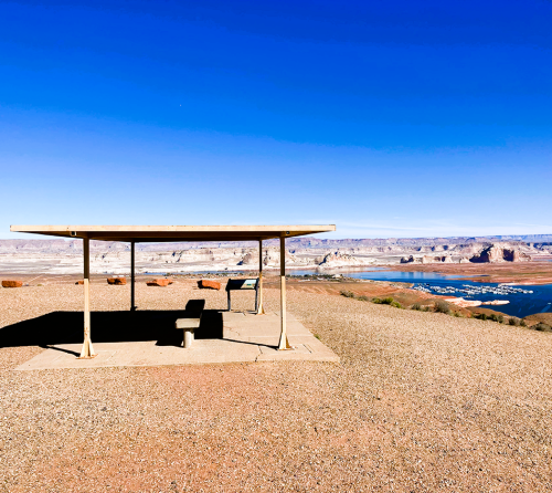 A shaded bench overlooks a scenic view of a lake and mountains under a clear blue sky.
