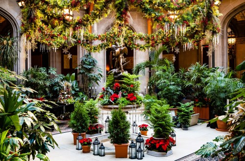 A festive indoor garden with lush plants, a fountain, and holiday decorations, including garlands and poinsettias.