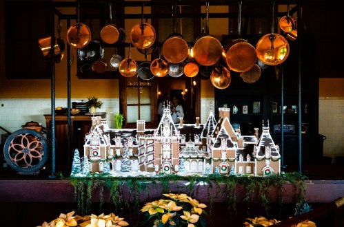 A festive display of a gingerbread house surrounded by holiday decorations and hanging copper pots in a cozy kitchen.