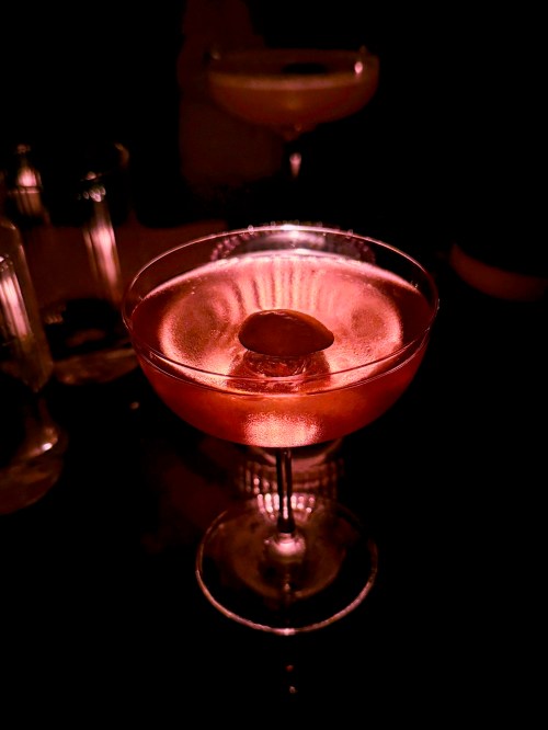 A close-up of a cocktail in a glass, illuminated softly against a dark background, with a cherry on top.