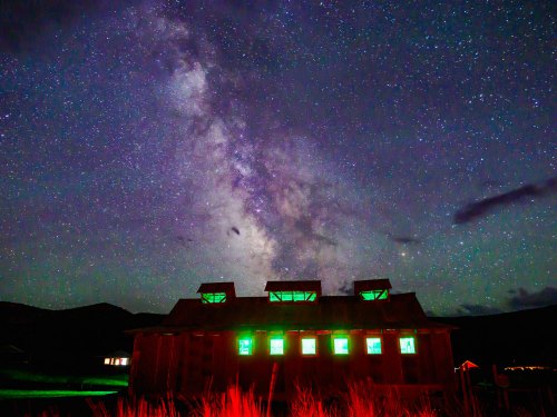 A starry night sky with the Milky Way above a rustic building illuminated by green lights.