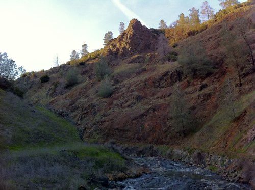 A rocky hillside with sparse trees, overlooking a small stream in a serene natural landscape.