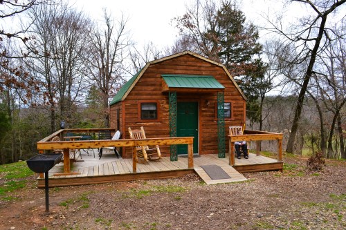 A rustic wooden cabin with a green roof, surrounded by trees and featuring a deck with chairs and a grill.